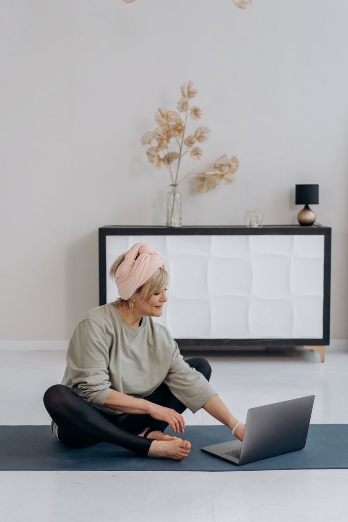 An older woman seated on a yoga mat and working on a laptop in a chic living room.