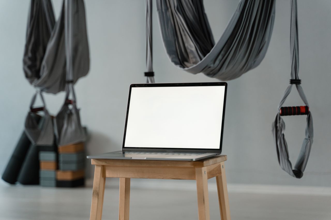 Studio de yoga épuré avec hamacs aériens et écran d’ordinateur portable vide posé sur un tabouret, idéal pour une variété de thèmes bien-être.